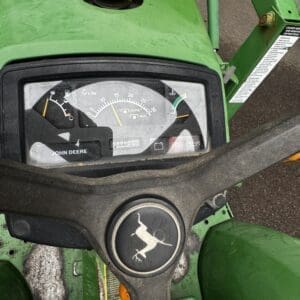 IMG_1394 - Montana Auctions by Kevin Hill Close-up of a green John Deere tractor dashboard with a circular gauge cluster and a black steering wheel bearing the leaping deer logo at center.