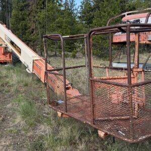 IMG_6541 - Montana Auctions by Kevin Hill Rusty orange industrial boom lift with a mesh-platform basket parked on grassy, dirt field near a forest edge.
