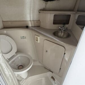 IMG_6565 - Montana Auctions by Kevin Hill Compact marine head with a toilet on the left, small sink, and cabinet; oval porthole above and beige surfaces showing dirt and wear.
