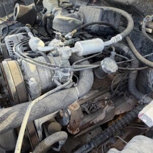 IMG_6702 - Montana Auctions by Kevin Hill Close-up of a dusty vehicle engine bay showing the carburetor, air intake, and various hoses and belts.