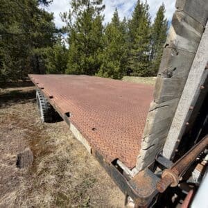 IMG_6830 - Montana Auctions by Kevin Hill Rusty diamond-plate flatbed trailer with a wooden side, parked among trees in a dry grassy area