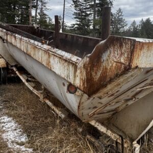 Rusty, abandoned dump trailer with peeling paint and heavy rust streaks sits on a snow-dlecked, grassy roadside among pine trees in a forested area.