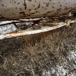 Rusty white cylindrical tank with brown rust spots and supporting pipes, siting in a field with dry grass and a light snow dusting.