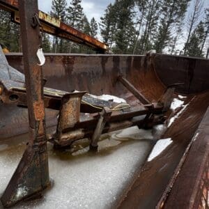 Rusty metal scoop and support frame inside a large rusted container, with ice forming on the bottom and trees in the background.