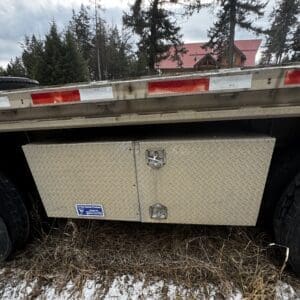 Diamond-plate storage toolbox mounted under a trailer, with red/white reflective bumper above and tires on either side.
