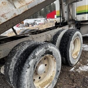 Rusty dump truck trailer with three axles and dirty, worn tires in a scrapyard yard under a raised bed.
