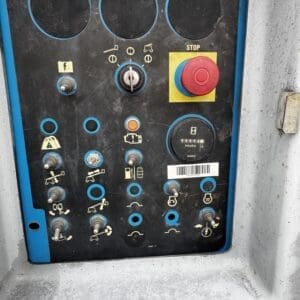 Industrial control panel with numerous switches and dials, a large red stop button, and an hour meter on a dusty dark panel framed in blue outer casing.