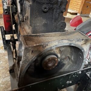 Rusty engine block mounted on a stand with a large rusted pulley, oil and dirt caked on the surfaces, outdoors in a shed.