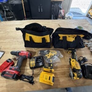 Two black-and-yellow tool bags sit on a workbench with cordless drills, batteries, and chargers laid out nearby in a workshop setting.