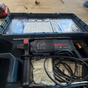 Open tool case containing a Bosch hammer drill with red trigger, a coiled cord, protective gloves, and an instruction sheet inside the lid.