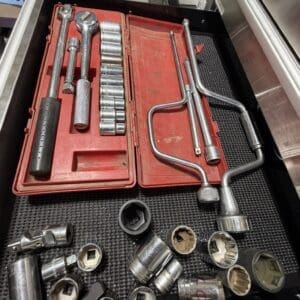 Red tool case opened to reveal sockets, ratchets, and extensions; loose socket pieces scattered on a black work mat nearby.