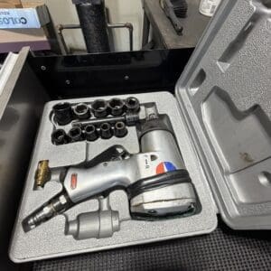 Air-powered impact wrench with multiple sockets arranged in a foam tray inside an open tool case on a workshop workbench.