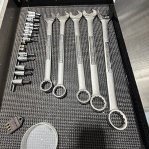 Six large Craftsman combination wrenches arranged in a row inside an open metal tool chest, with a row of sockets to the left and a small red-lit keypad device in the corner bottom left.