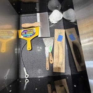 Tools laid out in a metal cabinet tray: yellow handheld scraper, wooden handles, metal blades, and sanding blocks/abrasives visible.