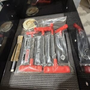 Set of small hand tools with red T-handle grips in sealed plastic packaging (Craftsman) lying in a vented metal tool drawer, with a wooden-handled brush nearby.