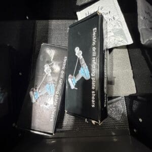 Two black boxes labeled 'Electric drill retaining plate shears' with an image of a blue cordless drill on the box, stored in a car trunk.