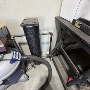 Tall rusty black cylinder connected to metal pipes in a cluttered workshop, with a flexible vacuum hose on the floor nearby.