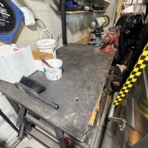 Cluttered metal workbench in a workshop, with a grinder on the right, a vise, a brush, paint bucket, and shelves of tools behind.