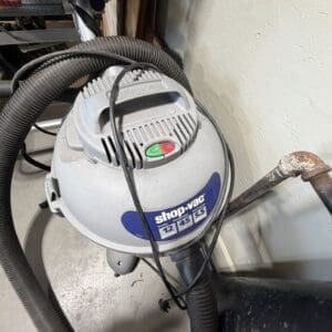 Wet/dry shop vacuum with hoses in a workshop, attached to a wall and near a rusty pipe edge.