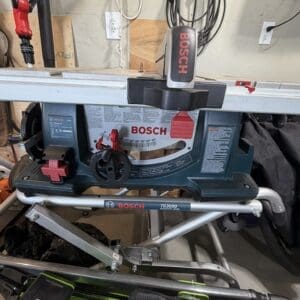 Bosch TS3000 miter saw on a metal stand in a cluttered workshop, cords hanging nearby and a red blade guard visible.
