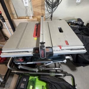 Bosch table saw on a stand in a cluttered workshop, with cords, wood scraps, and a green tool chest nearby.