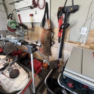 Cluttered home workshop with a miter saw on a stand, pegboard wall, and various power tools and cords on the bench and floor.