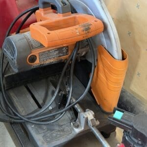 Orange and gray circular saw on a workshop stand with tangled cords and a red fuel can nearby.