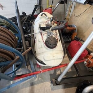 White plastic fuel tank on a cart surrounded by hoses, power tools, and red gas cans in a cluttered workshop.