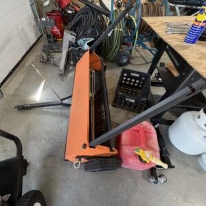 Orange metal wheeled frame or cart in a crowded workshop, with hoses, jugs, and tools scattered around.