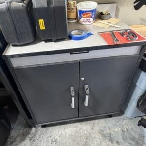 Two-door gray steel tool cabinet on wheels in a workshop, with clutter on top including a black tool case, a bottle labeled grease, a tub of dairy cheese, blue painter's tape, and a red number plate.