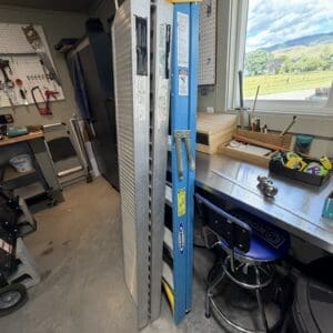 Workshop corner with several metal ladders leaning against a wall, a blue Werner ladder, and a pegboard with tools; a workbench and stool sit nearby beneath a window showing a green field and hills outside.