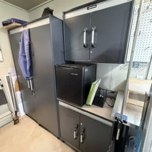 Gray metal garage/storage area with tall cabinet, overhead cabinet, and a small black mini-fridge on a workbench. A blue shop towel hangs from the tall cabinet; pegboard and tools are visible nearby.