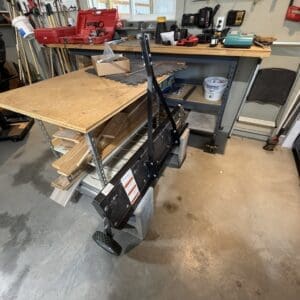 Cluttered wooden workbench in a workshop, with pegboard tools and a wheeled black metal cart in the foreground