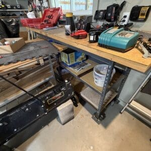 Workbench in a cluttered workshop with a red tool case, batteries, and a teal battery charger on a wooden bench under a pegboard wall.
