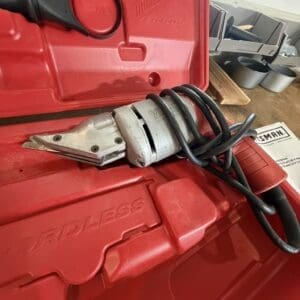 Red Milwaukee cordless tool case with a coiled black power cord and metal clamp/attachment resting on it, inside a workshop.