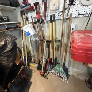Tools hanging on a pegboard wall in a cluttered garage/workshop: rakes, shovels, hoes, a bucket, and a red storage bin to the right.