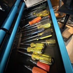 Assorted screwdrivers with yellow and orange handles laid out inside a blue tool chest drawer in a workshop.