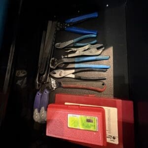 Open tool drawer with assorted hand tools: blue-handled pliers, needle-nose pliers, files, and a red plastic toolbox case.