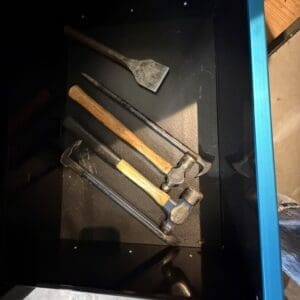 Assorted hand tools (hammers, a chisel, and a bar) laid in a black toolbox drawer with a blue frame edge
