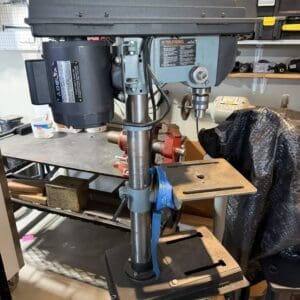 Bench-mounted drill press on a stand in a workshop, with a pegboard and tools visible in the background.