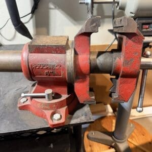Red Roughneck bench vise gripping a metal shaft on a workbench; a grinding wheel is mounted on a spindle in the background.