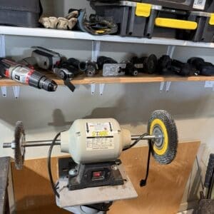 Bench grinder with two circular buffing wheels on a stand, in a workshop with tool storage above.