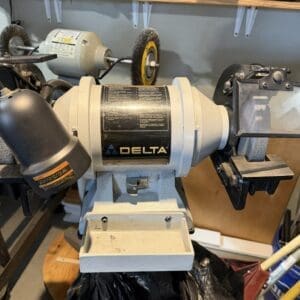 Delta bench grinder on a metal stand in a workshop, with two grinding wheels and a cluttered workbench behind it.
