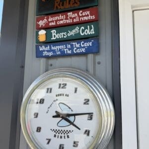 Colorful wooden signs say 'MAN CAVE Rules', 'Beers always Cold', and 'What happens in The Cave stays in The Cave', mounted above a round chrome-diner clock.