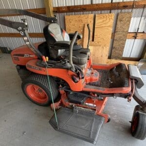 Red Kubota ZD1011 riding lawn mower parked in a shed, with a front attachment and wooden boards on the wall behind it.