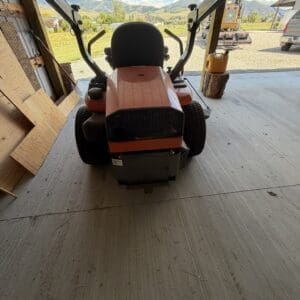 Orange riding lawn mower inside a shed, facing toward a rural yard and distant mountains.