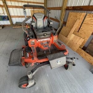 Red riding lawn mower with a black seat and roll bar parked inside a wooden-shed workspace, front cutting deck visible.
