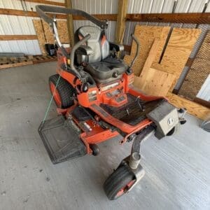 Red riding lawn mower in a shed with plywood walls and concrete floor, front wheels and a grass catcher attached at the back side rail bar nearby