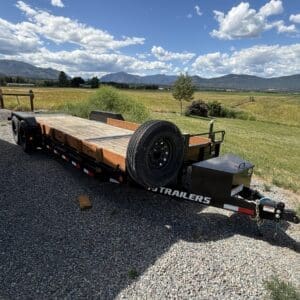 Flatbed trailer with a spare tire mounted on the front, parked on gravel with open field and mountains in the distance under a blue sky.