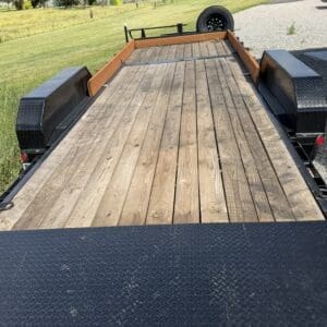 Flatbed trailer with wooden deck and diamond-plate fenders parked on a grassy rural area with mountains in the background